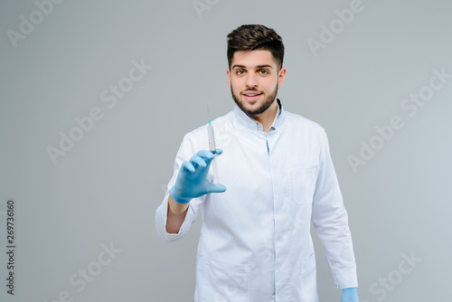 Handsome medical doctor in gloves with suringe smiling isolated over grey background photo