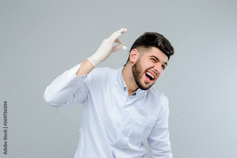 Young medical doctor in gloves treating his ears with drops isolated over grey background Young medical doctor in gloves treating his ears with drops isolated over grey background