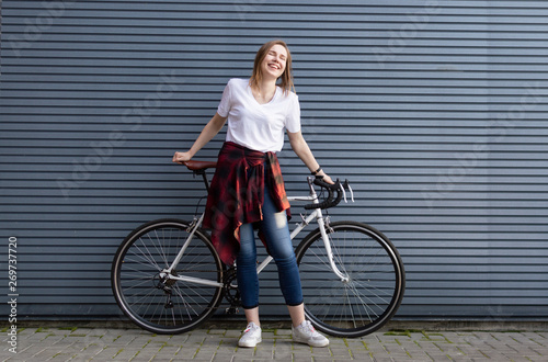 beautiful young girl standing with a white bicycle on a background of gray striped wall, the woman is happy