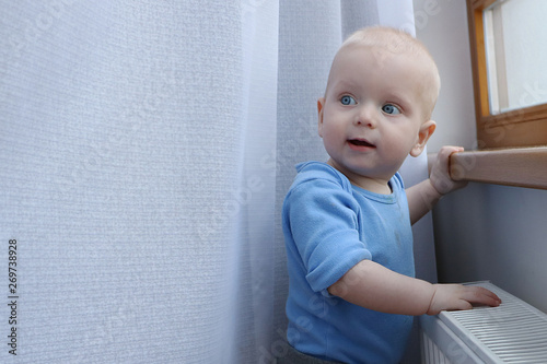 One year old baby boy with blue eyes is playing with curtains near window. Need to be careful with choosing children playground