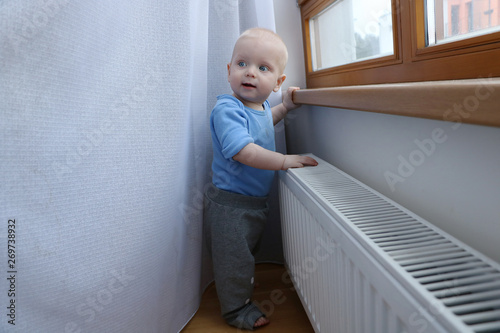 One year old baby boy with blue eyes is playing with curtains near window. Need to be careful with choosing children playground