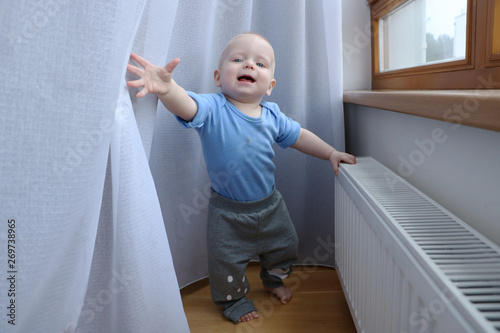 One year old baby boy with blue eyes is playing with curtains near window. Need to be careful with choosing children playground