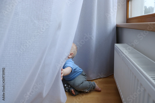 One year old baby boy with blue eyes is playing with curtains near window. Need to be careful with choosing children playground