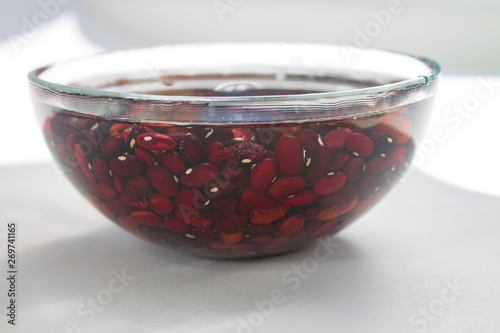 lots of raw red bean beans soaked in water in a clear glass bowl on a white table with sun shade