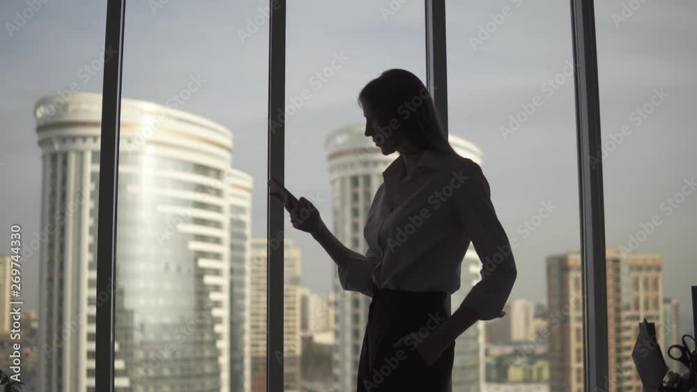 Silhouette of a young woman against the background of a large window and skyscraper. girl uses a smartphone.