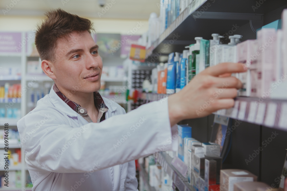 Cheerful handsome male pharmacist doing stocktaking, organizing products on shelves at drugstore. Charming young male chemist working at the pharmacy. Retail, selling medical goods concept
