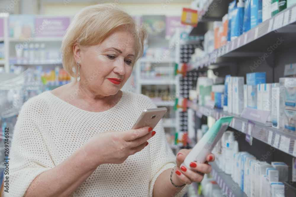 Senior woman using her smart phone at the drugstore, scanning qr code of a product. Elderly female customer browsing online description of a medical product, shopping at pharmacy