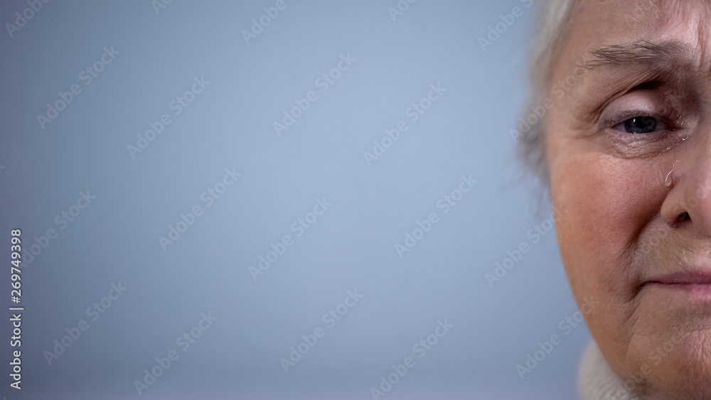 Fototapeta premium Half-face of upset crying elderly woman on grey background, poverty hopelessness