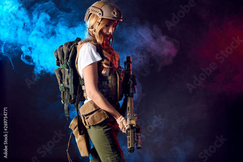 Female storm trooper in military camouflage uniform protected with helmet, body armour with submachine gun seeking aims in the smoky darkness