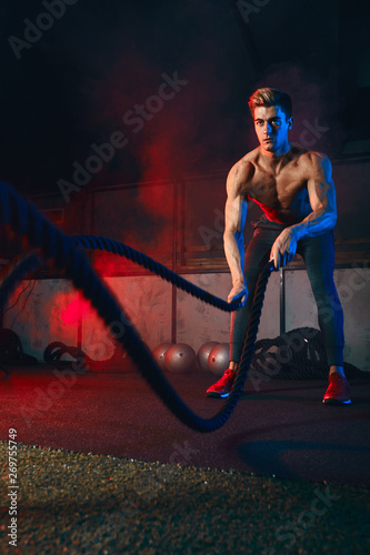 Determined shirtless male athlete performing waves by battle rope, keeping fit in cross fit club.