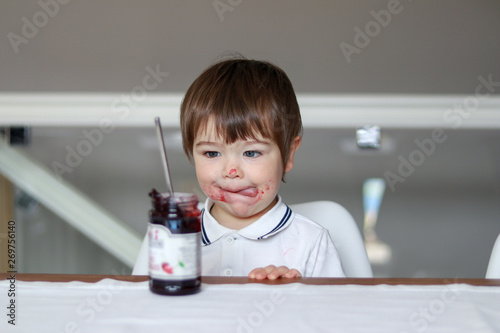 Portrait of funny little boy looking at glass jar with cherry jam with his tongue out and dirty face. Sweet tooth. Child nutrition concept.