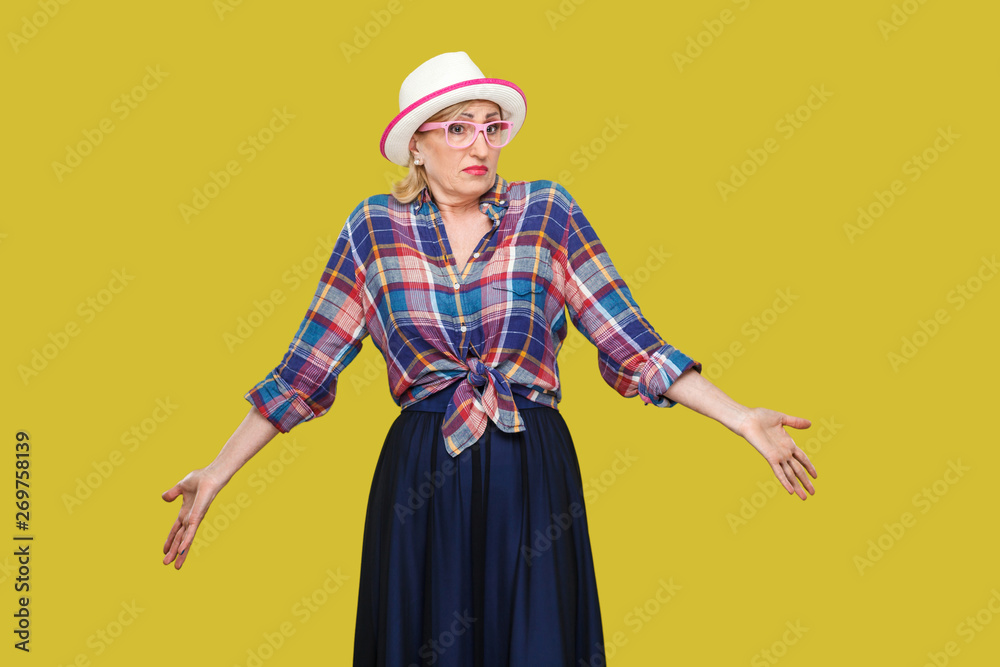 I don't know. Portrait of confused modern stylish mature woman in casual style with hat and eyeglasses standing, thinking, raised arms, looking away. indoor studio shot isolated on yellow background.