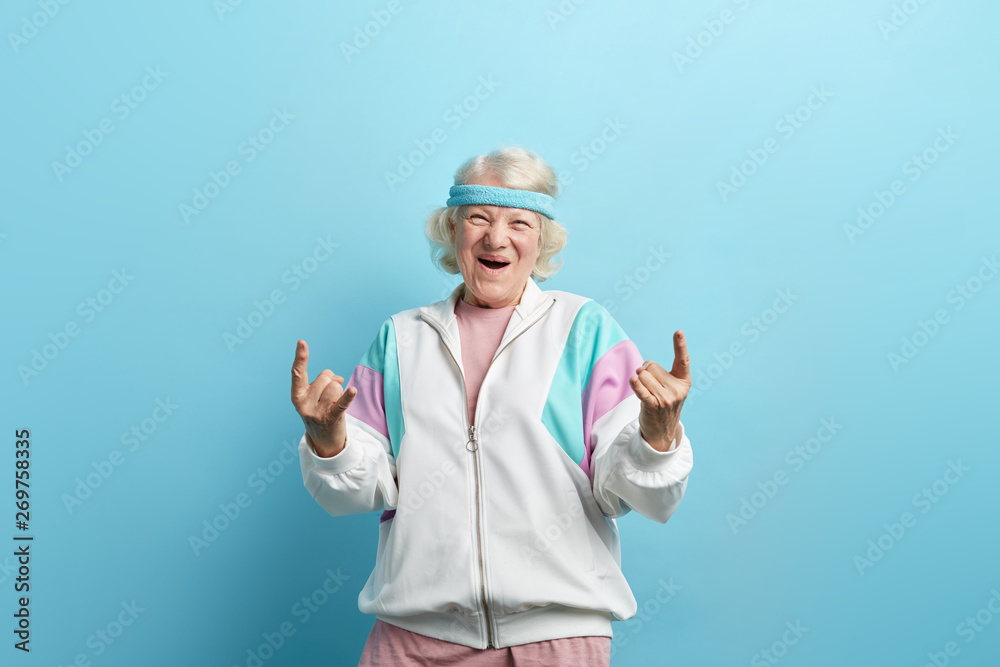 Cute hipster grandmother smiling and making rock sign against blue studio background - obrazy, fototapety, plakaty 