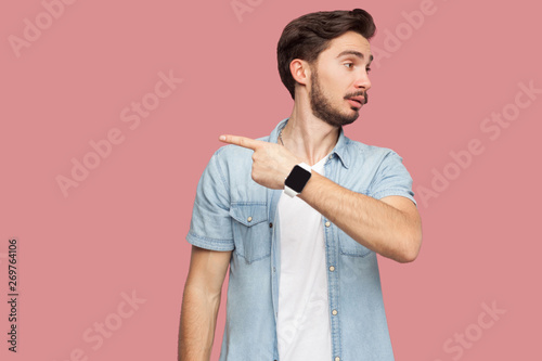 Get out of here. Portrait of angry handsome bearded young man in blue casual style shirt standing and showing out side exit way. indoor studio shot, isolated on pink background.