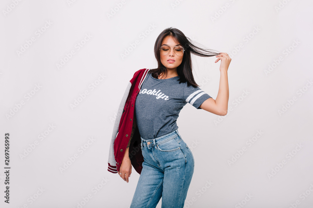 Shapely dark-haired girl sensually posing in studio holding stylish jacket on shoulder. Spectacular brunette female model in jeans dancing with eyes closed in front of white wall.