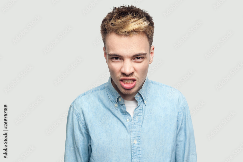Portrait of angry aggressive handsome young man in light blue shirt standing and looking at camera with bad mood, clenching teeth and angry. indoor studio shot on grey background copy space. Portrait of angry aggressive handsome young man in light blue shirt standing and looking at camera with bad mood, clenching teeth and angry. indoor studio shot on grey background copy space.