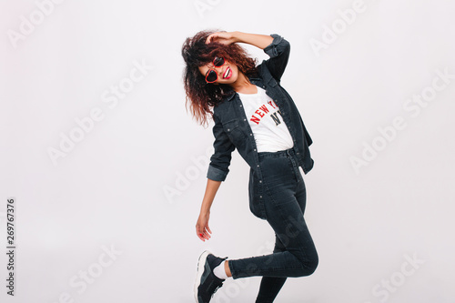 Amazing slim female model in black pants standing on one leg isolated on white background with smile. Curly brunette girl dancing in trendy sneakers and smiling.
