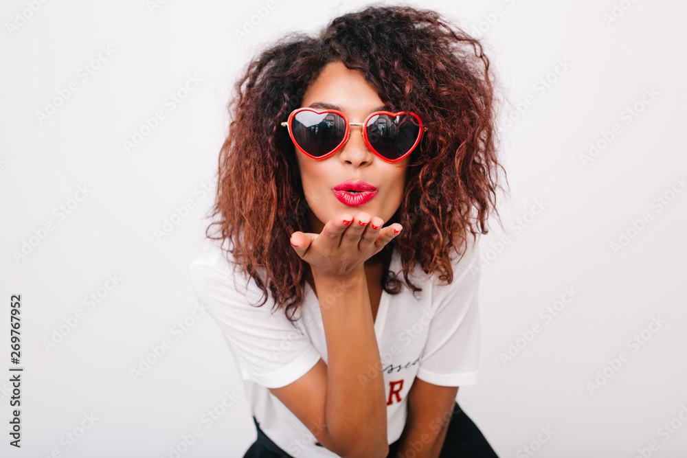 Lovely lady with light-brown skin sending air kiss isolated on white background. Beautiful african female model with curly hairstyle posing in trendy red sunglasses in studio.