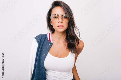 Close-up portrait of stunning dark-haired girl in white tank-top looking with interest to camera. Indoor photo of adorable european lady in stylish glasses chilling on photoshoot.