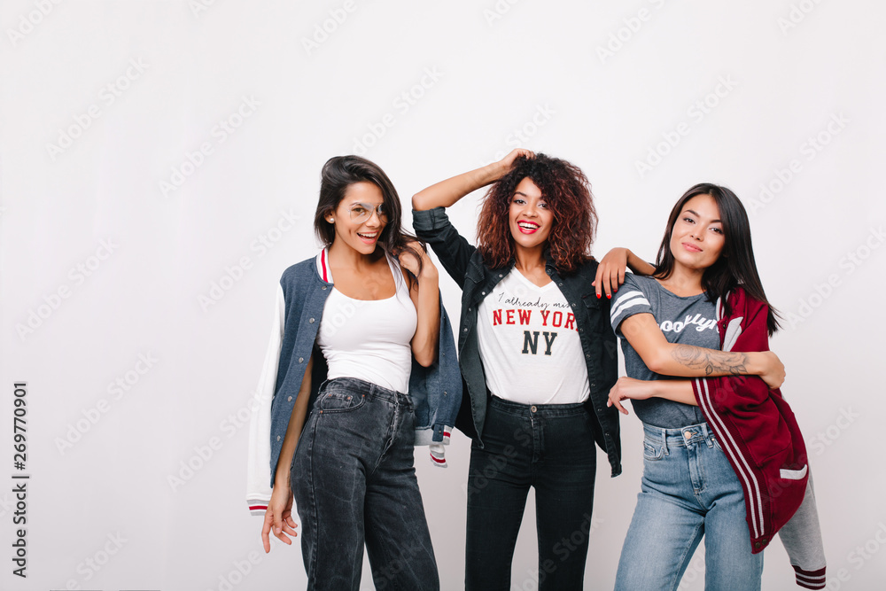 Obraz premium Blissful latin girl in jeans having fun with university mates posing in studio with smile. Indoor portrait of lovely african lady standing between friends and playing with curly hair.