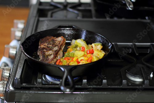New York strip steak frying in a cast iron pan with potatoes,assorted bell peppers, and onions on the stove top.