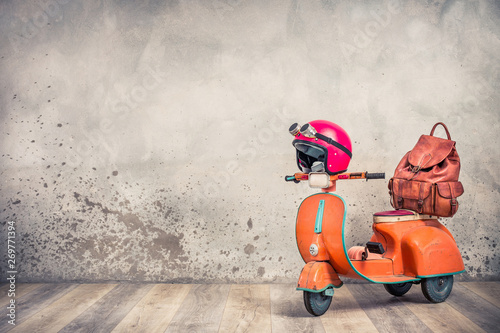 Retro orange toy old rusty children's pedal scooter from 60s, red helmet with aviator goggles and leather backpack front loft concrete wall background. Travel concept. Vintage style filtered photo