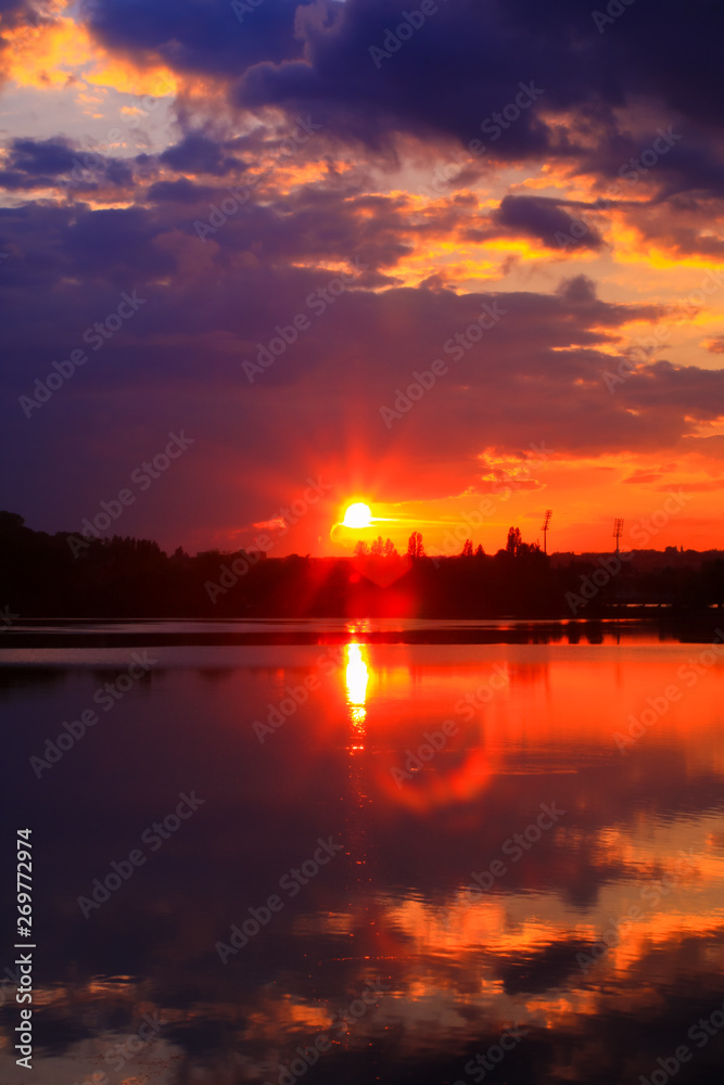 Vertical sunset on the water with symmetry of colorful clouds cumulonimbus. Golden orange reflection of the sun's rays in a lake. Lens flare in foreground Vertical sunset on the water with symmetry of colorful clouds cumulonimbus. Golden orange reflection of the sun's rays in a lake. Lens flare in foreground