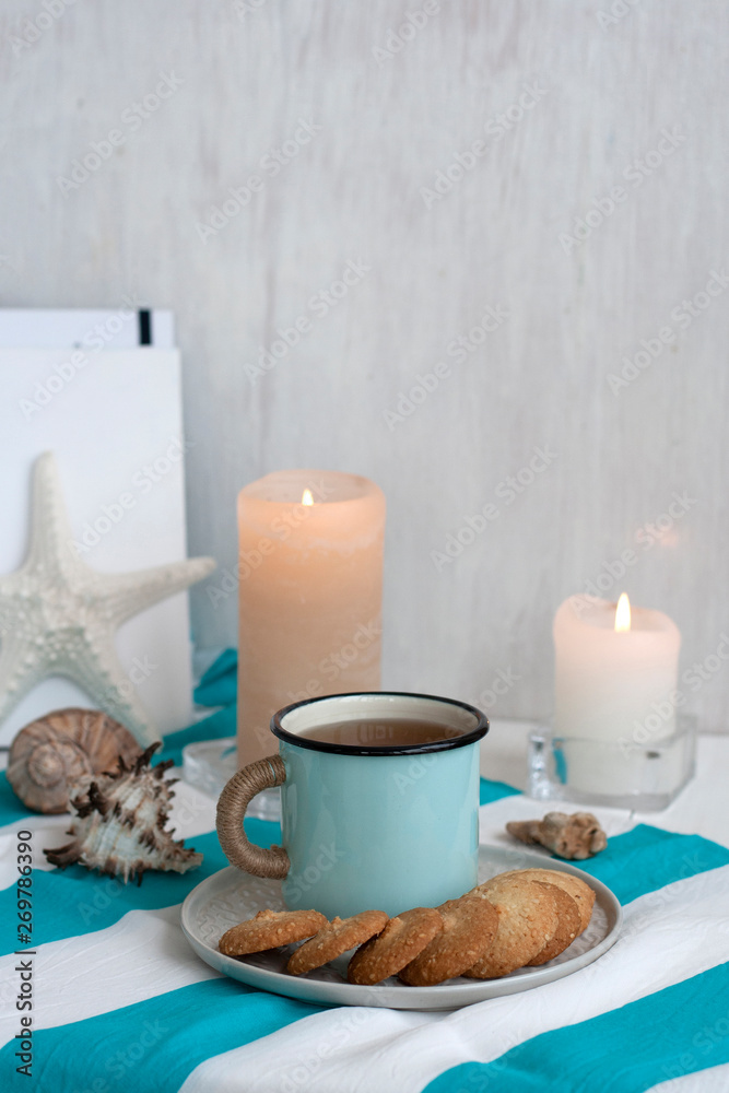 A hot mug of black tea with homemade cookies on a striped tablecloth, wax candles, decorative starfish, seashell, pile of notebooks on a white background.