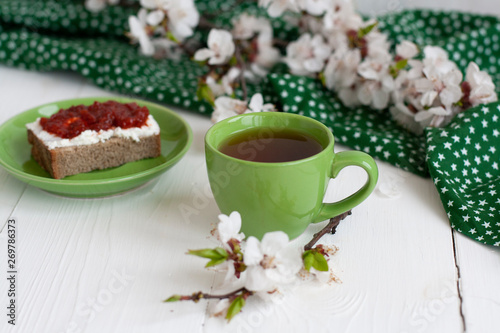 A cup of black tea on a green napkin, homemade rye bread with soft cream cheese and smoky dried tomatoes, a branch of apricot bloom on the white background.