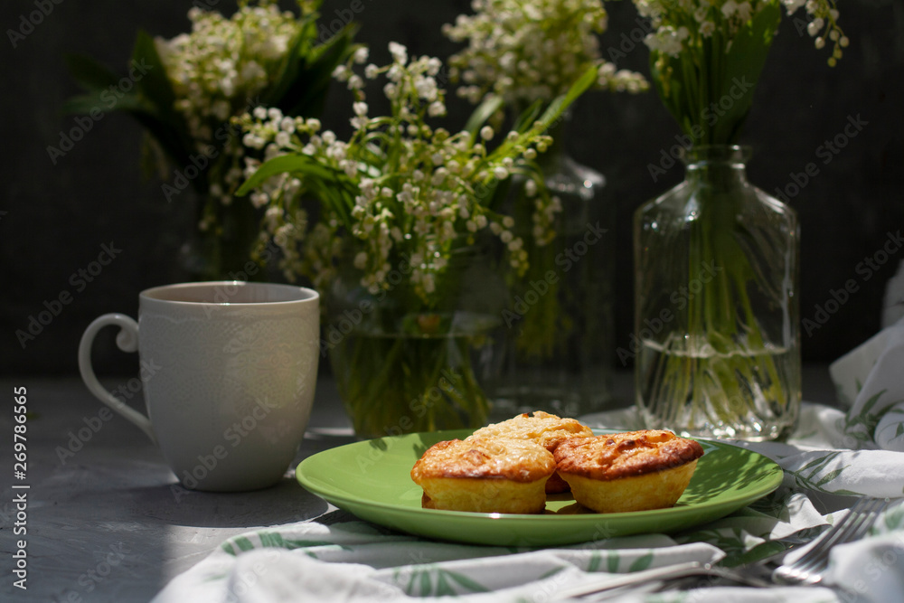A tasty breaksfast. A plate of freshly-baked muffins with a hot cup of coffee on a white tablecloth, bouquets of lilies of the valley in glass vases on the gray background. A tasty breaksfast. A plate of freshly-baked muffins with a hot cup of coffee on a white tablecloth, bouquets of lilies of the valley in glass vases on the gray background.