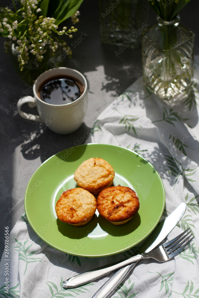A tasty breaksfast. A plate of freshly-baked muffins with a hot cup of coffee on a white tablecloth, bouquets of lilies of the valley in glass vases on the gray background.