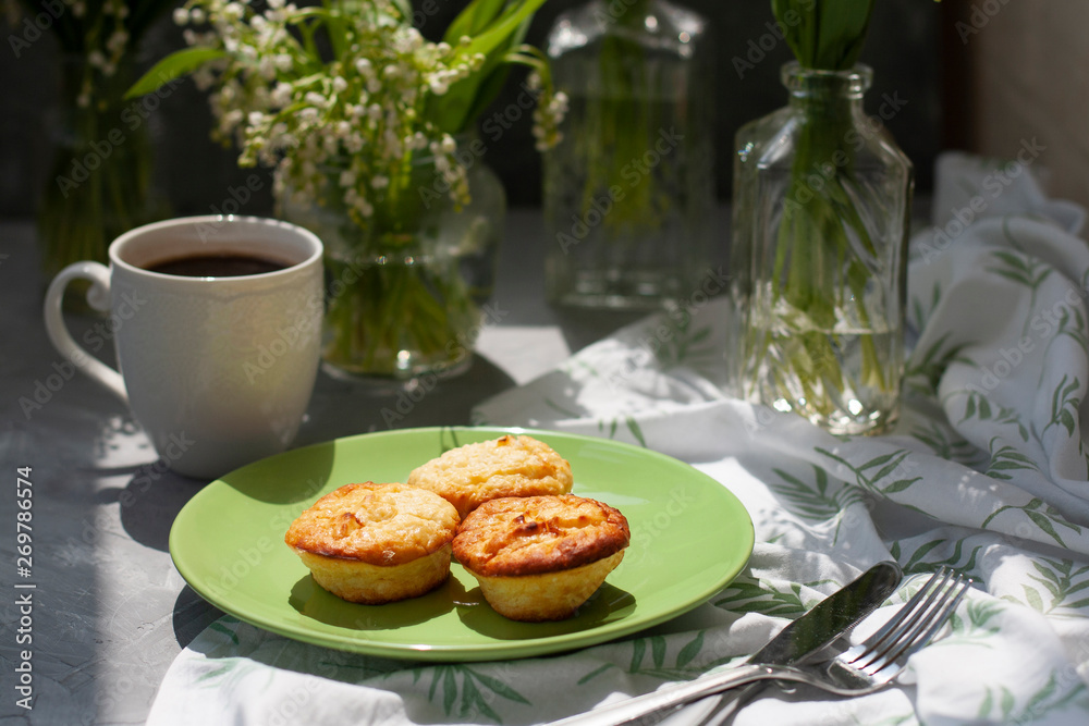 A tasty breaksfast. A plate of freshly-baked muffins with a hot cup of coffee on a white tablecloth, bouquets of lilies of the valley in glass vases on the gray background. A tasty breaksfast. A plate of freshly-baked muffins with a hot cup of coffee on a white tablecloth, bouquets of lilies of the valley in glass vases on the gray background.