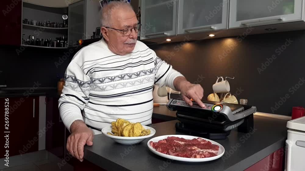 serious senior man in white striped sweater switches on and prepares electric grill in modern kitchen close view serious senior man in white striped sweater switches on and prepares electric grill in modern kitchen close view