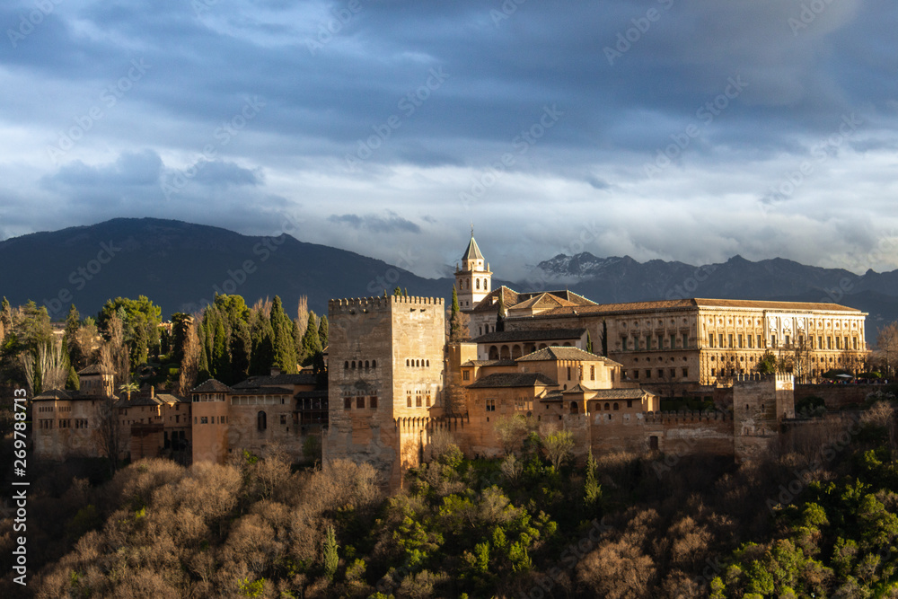 La Alhambra lit by the sunset in Granada under a cloudy sky.