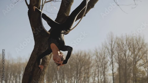 Beautiful woman doing some acrobatic elements on aerial hoop outdoors. Flexible brunette with bare feet hanging in ring for aerial acrobatics. Medium shot