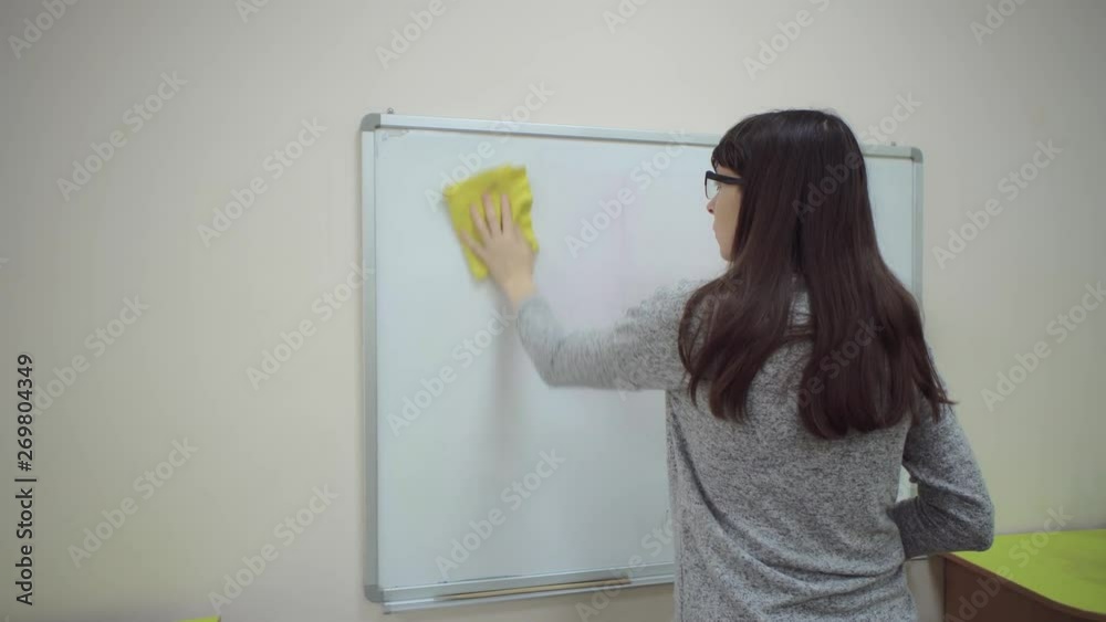 Female teacher stands at whiteboard and explains rules of addition in elementary school. Caucasian schoolmaster in glasses removes the examples with a board duster. Female teacher stands at whiteboard and explains rules of addition in elementary school. Caucasian schoolmaster in glasses removes the examples with a board duster.