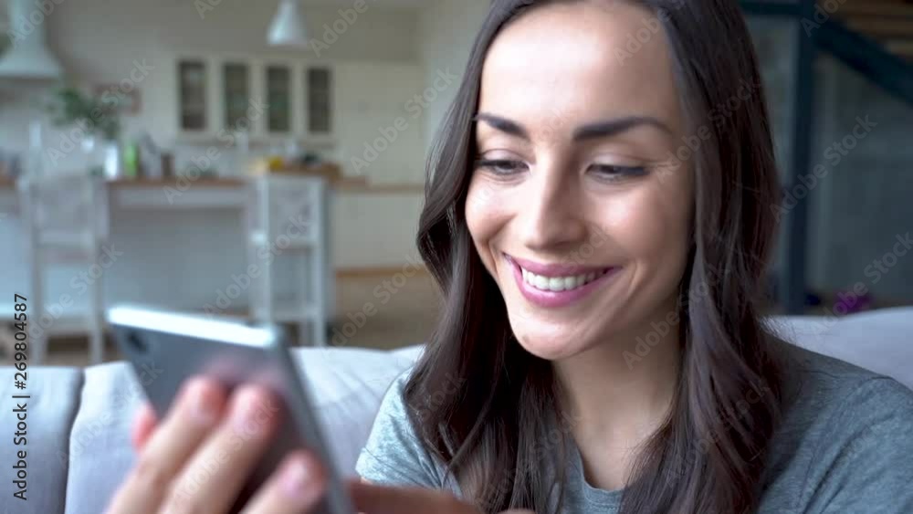 Slow motion video of happy beautiful young woman while she using mobile phone and sitting on the couch at home is surfing in internet or chatting in social network. Slow motion video of happy beautiful young woman while she using mobile phone and sitting on the couch at home is surfing in internet or chatting in social network.