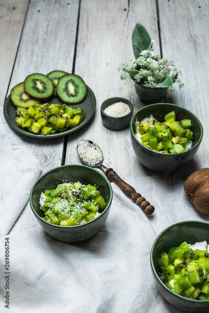 on a white, wooden table top, green tableware with fresh yoghurt and pieces of kiwi fruit sprinkled with coconut flakes, next to a tiny bouquet of field flowers and leaves in green and white color on a white, wooden table top, green tableware with fresh yoghurt and pieces of kiwi fruit sprinkled with coconut flakes, next to a tiny bouquet of field flowers and leaves in green and white color