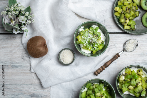 on a white, wooden table top, green tableware with fresh yoghurt and pieces of kiwi fruit sprinkled with coconut flakes, next to a tiny bouquet of field flowers and leaves in green and white color