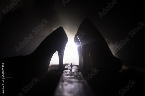 Artwork decoration. Silhouette of a man standing in the middle of the road on a misty night with giant high heel women shoes. Women power or women domination concept.