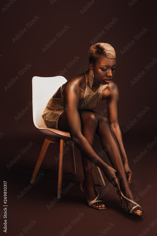 Naklejka premium fashionable african american woman with short hair sitting on chair on brown