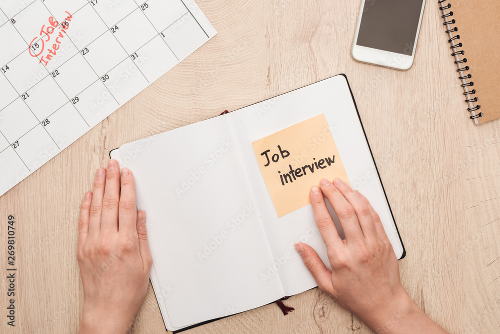 Obraz premium cropped view of recruiter holding notebook with job interview lettering written on sticky note