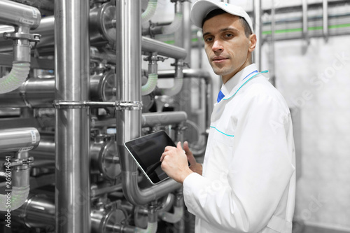 Portrait of man in a white robe and a cap standing in production department of dairy factory with grey tablet