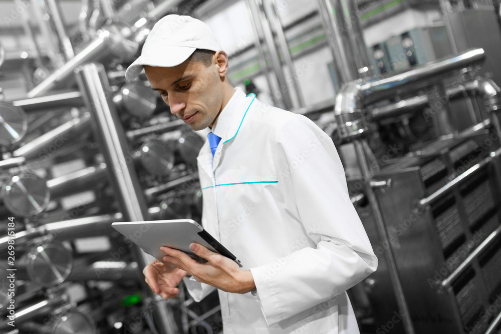 Portrait of man in a white robe and a cap standing in production department of dairy factory with grey tablet Portrait of man in a white robe and a cap standing in production department of dairy factory with grey tablet