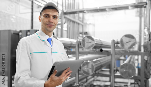 Portrait of man in a white robe and a cap standing in production department of dairy factory with grey tablet