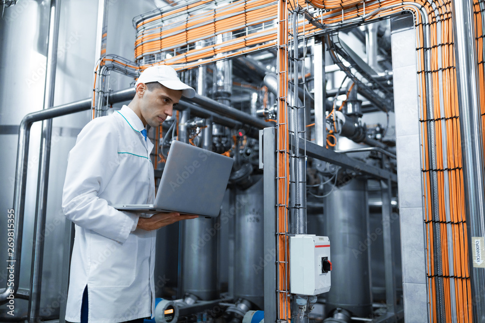 Portrait of man in a white robe and a cap standing in production department of dairy factory with laptop Portrait of man in a white robe and a cap standing in production department of dairy factory with laptop