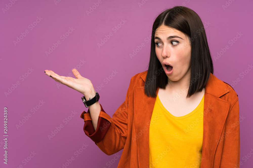 Young woman with a coat over isolated purple background holding copyspace imaginary on the palm Young woman with a coat over isolated purple background holding copyspace imaginary on the palm