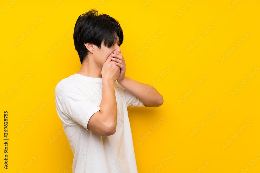 Asian man over isolated yellow wall covering mouth and looking to the side Asian man over isolated yellow wall covering mouth and looking to the side