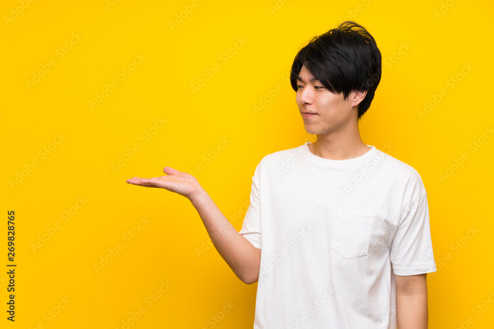 Asian man over isolated yellow wall holding copyspace with doubts Asian man over isolated yellow wall holding copyspace with doubts
