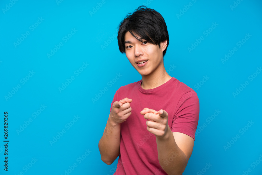 Asian man with red shirt over isolated blue wall points finger at you Asian man with red shirt over isolated blue wall points finger at you